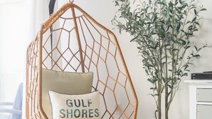 Una silla colgante de mimbre con un cojín que dice "GULF SHORES ALABAMA, USA" en una habitación con una planta en maceta y un armario blanco.