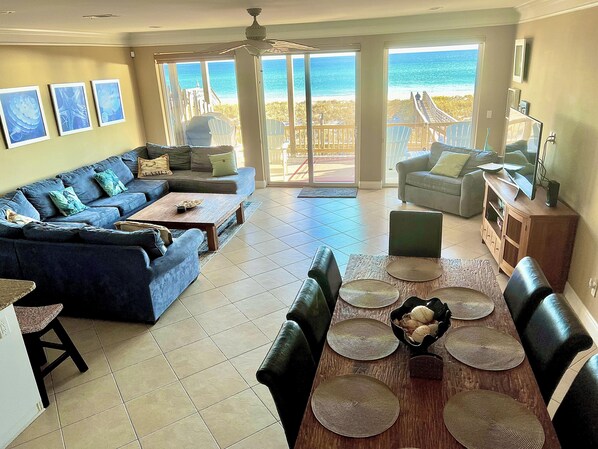 A spacious living room with a large sofa, a dining table with placemats, and a view of the ocean through large sliding glass doors.