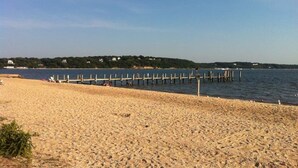 Une plage avec une jetée en bois qui s’avance dans des eaux calmes, un rivage sablonneux et une ligne côtière lointaine parsemée de bâtiments.