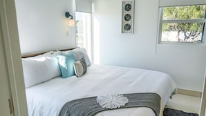 Ein ordentlich bezogenes Bett mit weißer Bettwäsche und einer grauen Decke, ein Fenster mit Blick auf Bäume und eine wandmontierte Lampe.