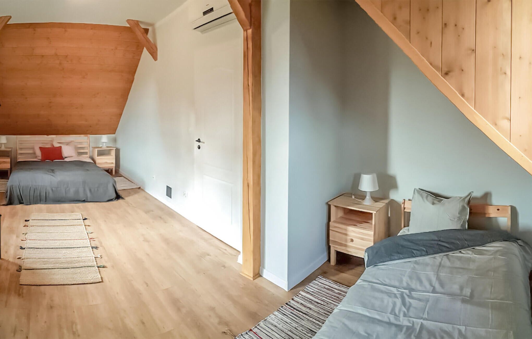 Una camera da letto con soffitto a falde, travi in legno, un letto con copripiumino grigio, un comodino con una lampada e un piccolo mobiletto in legno.