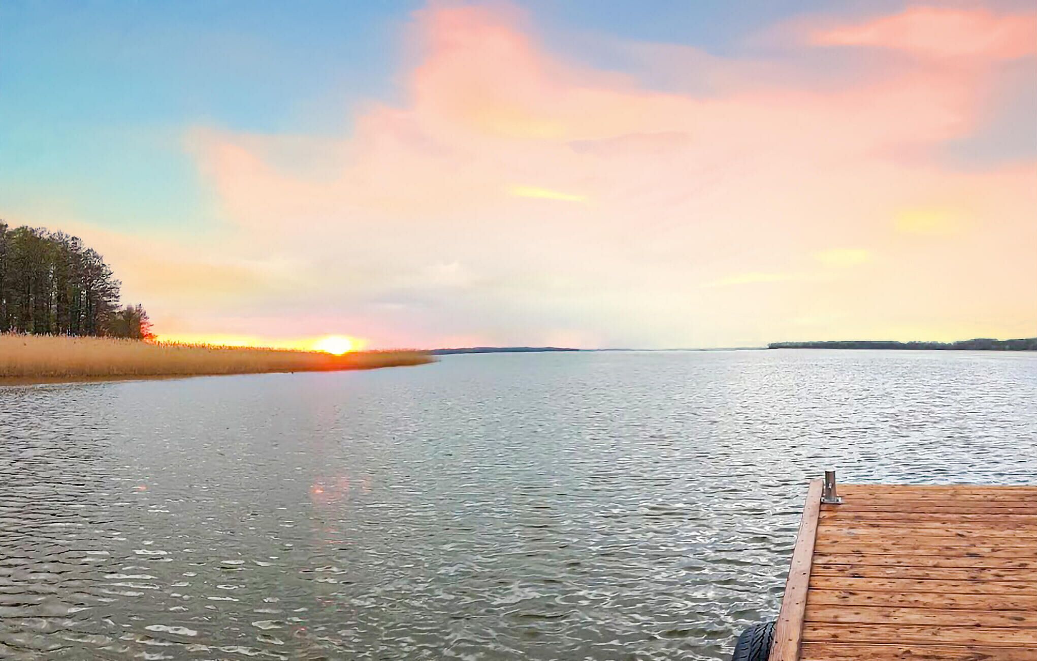 Ein ruhiger See mit einem hölzernen Steg, der ins Wasser ragt, umgeben von Bäumen und einem Sonnenuntergang im Hintergrund.
