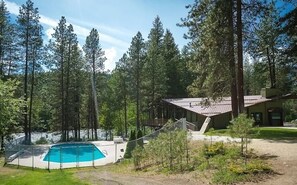 A house with a swimming pool surrounded by tall trees, a wooden deck, and a fenced area.