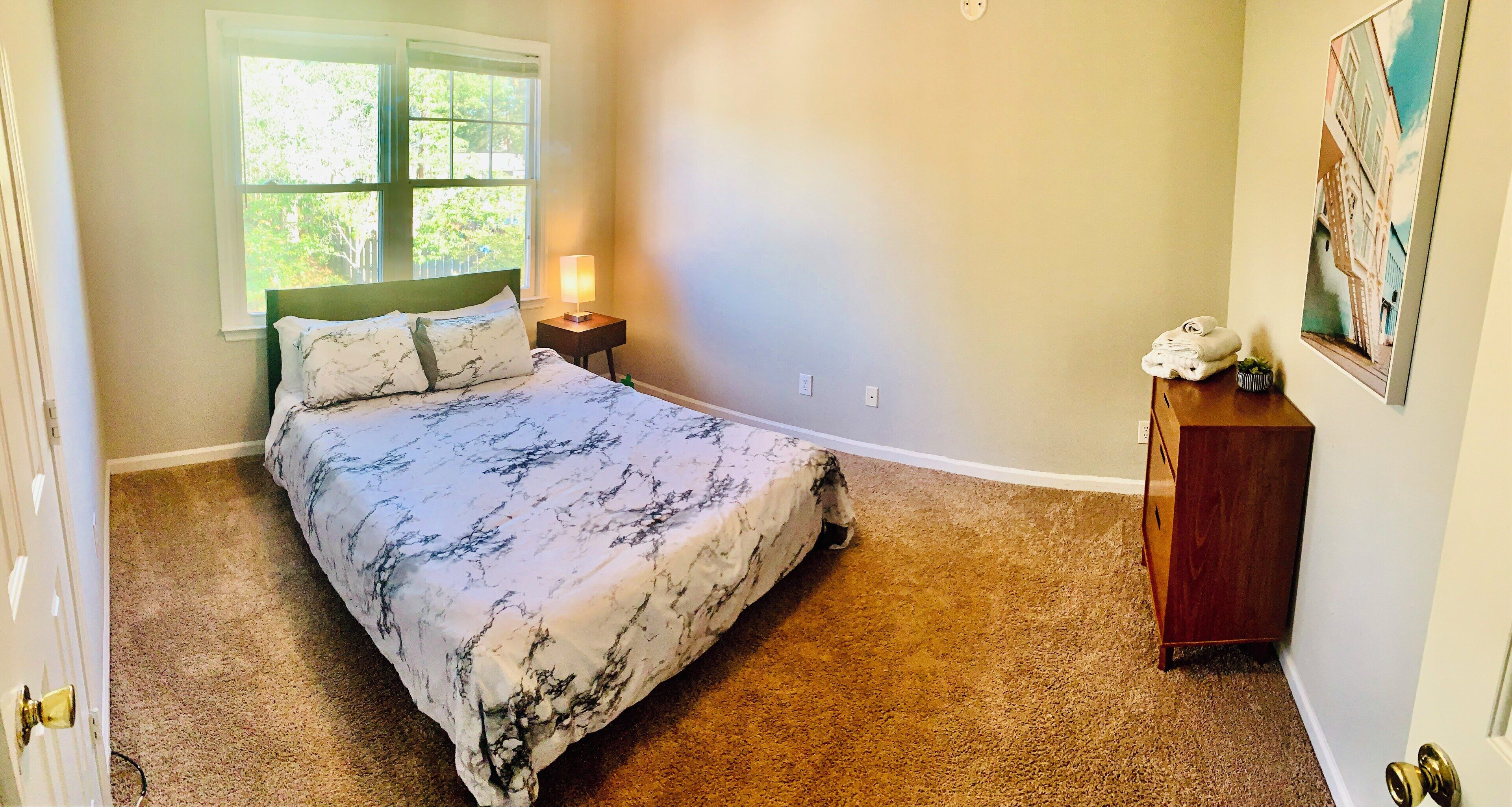 Un dormitorio con cama, mesita de noche, cómoda, ventana y un cuadro en la pared.