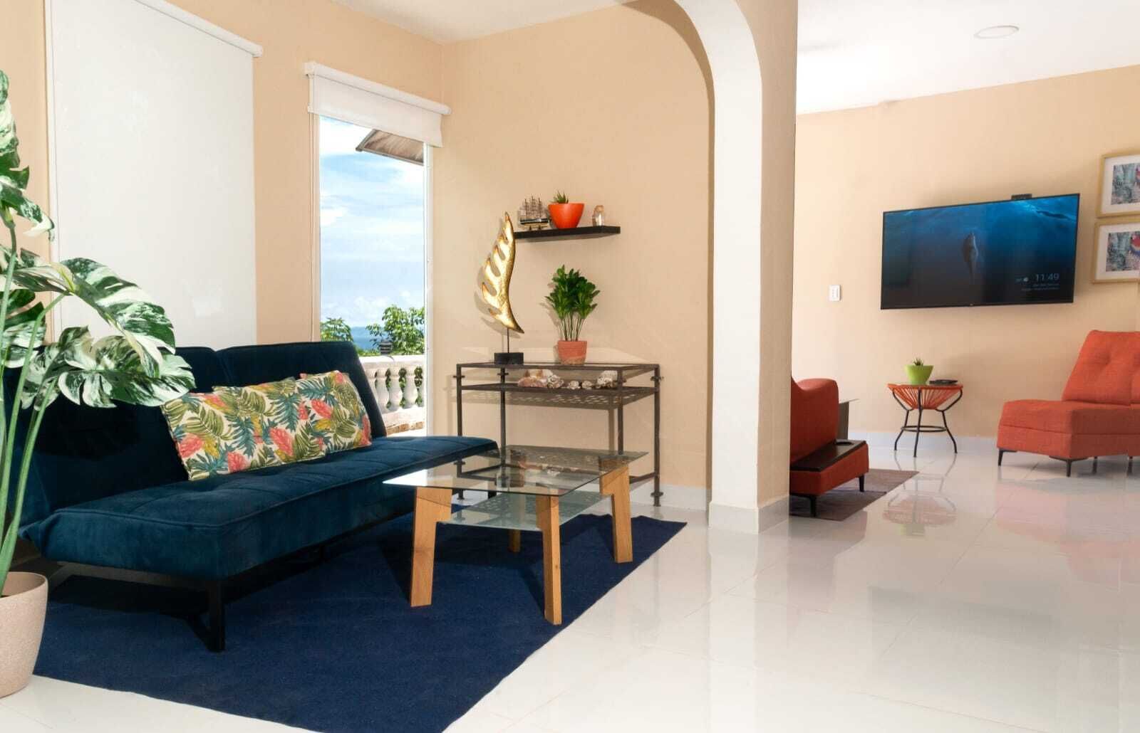 A modern living room with a blue sofa, a glass coffee table, a small side table, a red armchair, and a TV mounted on the wall.