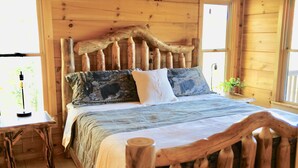 Camera da letto in una baita di legno con un letto caratterizzato da una testiera particolare, un comodino e una finestra vista verde.
