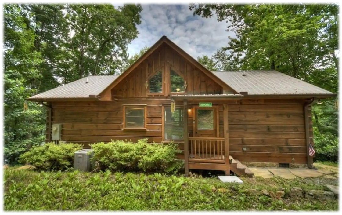 A wooden cabin with a gabled roof, a front porch, and a small front yard.