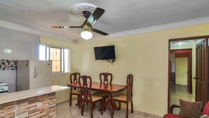 A dining area with a wooden table and chairs, a ceiling fan, and a TV mounted on the wall. - Family 1bedroom Apartment 15 Minute to the Airport (Santo Domingo Este)
