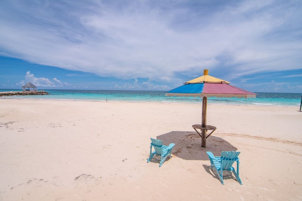 A beach with a colorful umbrella, two turquoise chairs, and a small hut in the background.