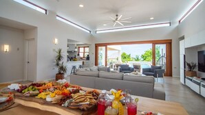 Ein geräumiges Wohnzimmer mit einem großen Holztisch, der für eine Mahlzeit gedeckt ist, einem grauen Sofa und einem Fernseher auf der rechten Seite.