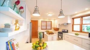 Eine moderne Küche mit weißen Schränken, einer zentralen Kochinsel mit Obst, Pendelleuchten und einem Fenster mit Jalousien.