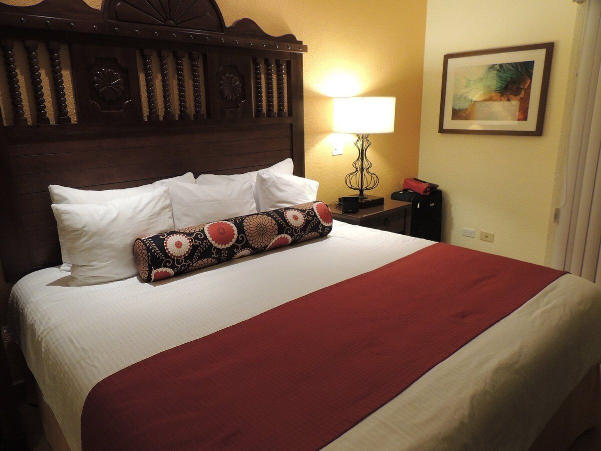 A neatly made bed with white and red bedding, a decorative pillow, a wooden headboard, a bedside lamp, and a framed picture on the wall.