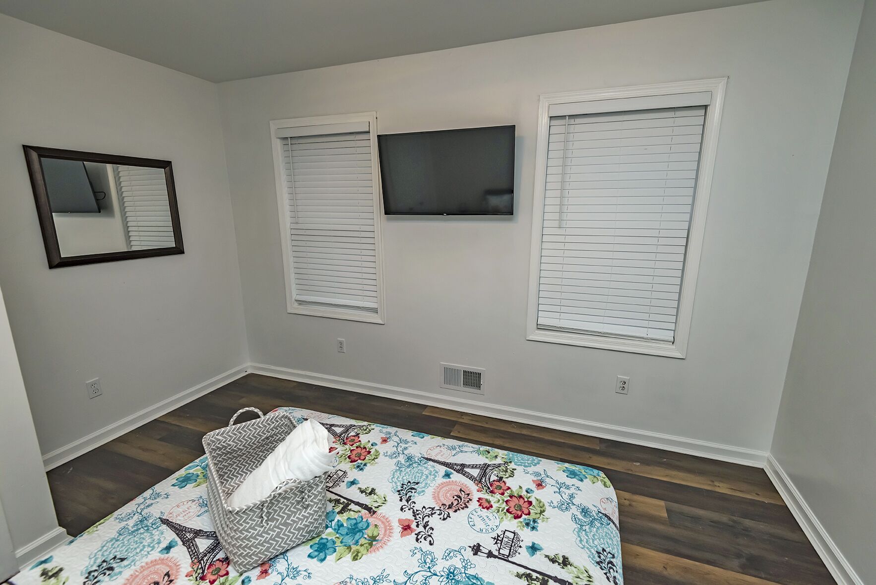 A room with a bed, a TV mounted on the wall, and two windows with blinds.