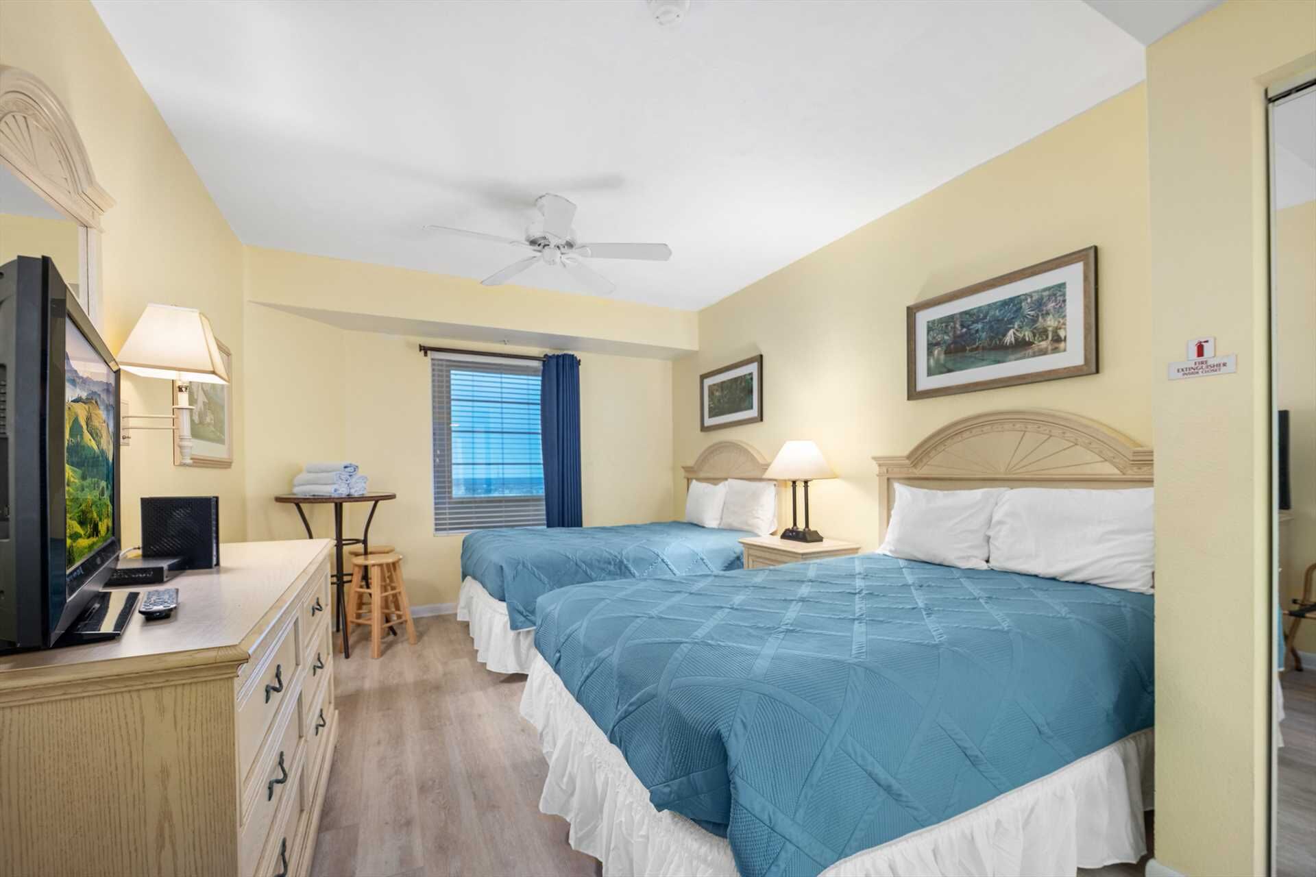 A hotel room with two beds, a desk with a television, a small table with towels, and a window with curtains.