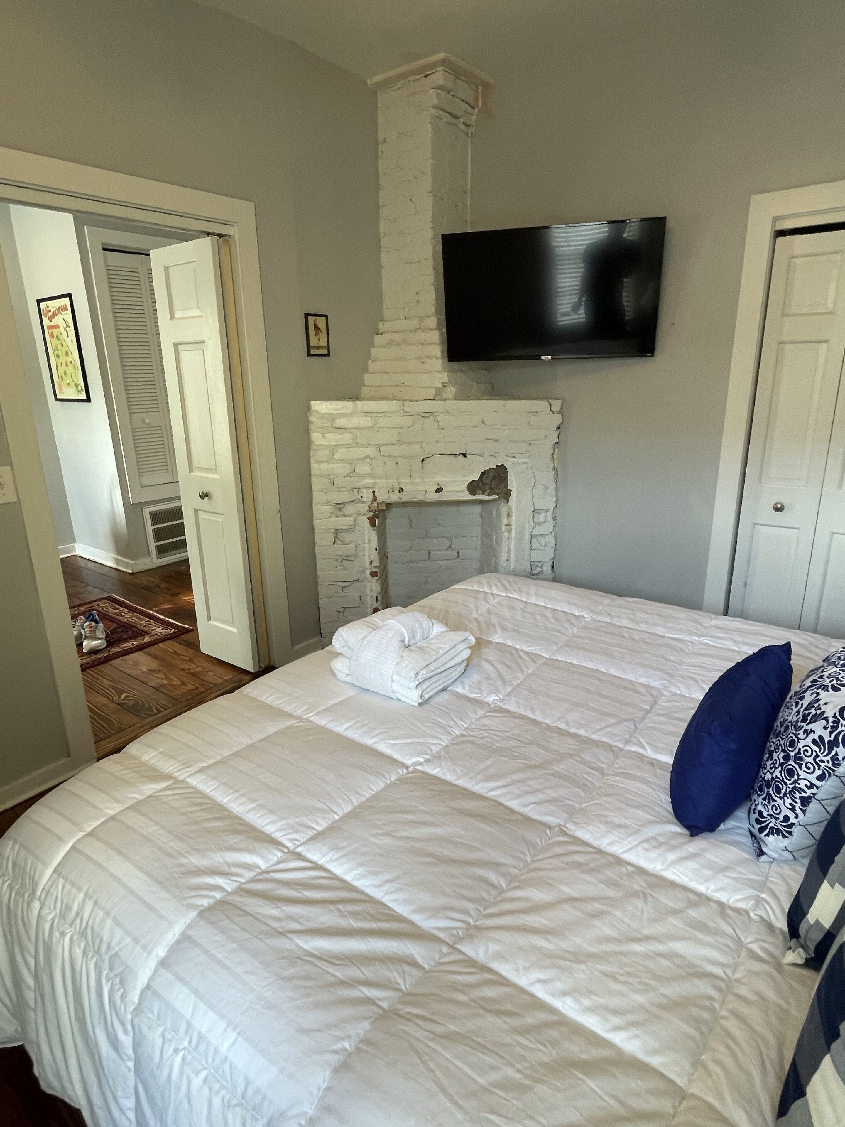 A bedroom with a white brick fireplace, a flat-screen TV mounted above it, and a neatly made bed with white and blue pillows.