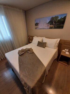 A neatly made bed with a brown blanket and pillows, a wooden headboard, a nightstand with a lamp, and a painting of a landscape on the wall. - Hotel (Gramado)