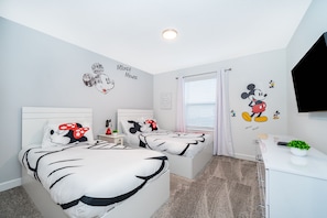 Quarto com duas camas, paredes brancas, decorações do Mickey Mouse, uma escrivaninha branca e uma televisão.