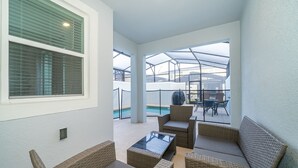 Eine überdachte Terrasse mit Korbmöbeln, einem Glastisch und Blick auf einen Swimmingpool.