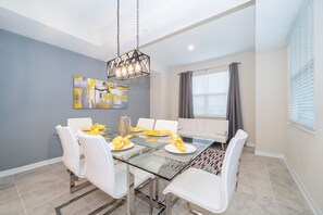 Uma área de jantar com mesa de vidro, cadeiras brancas, luminária pendente moderna e uma pintura contemporânea na parede.