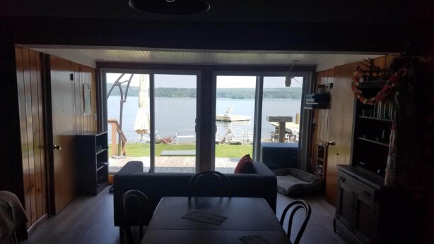 A room with a large sliding glass door leading to a lakeside deck. Inside, there is a dining table with chairs, a sofa, and a small kitchen area with a refrigerator and shelves. - Rustic-chic cottage with fantastic lake access, 15 minutes to Ithaca (Ithaca)