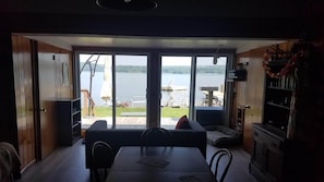 A room with a large sliding glass door leading to a lakeside deck. Inside, there is a dining table with chairs, a sofa, and a small kitchen area with a refrigerator and shelves.