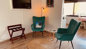 Ein Wohnzimmer mit an der Wand montiertem Fernseher, einem Tahiti-Poster und zwei türkisfarbenen Sesseln mit einem kleinen Tisch und einem gewebten Teppich.