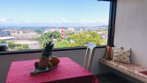 Ein Balkon mit Tisch, bedeckt mit einer roten Tischdecke, einem Stuhl und einer Bank mit Kissen. Auf dem Tisch liegen eine Ananas und Orangen. Im Hintergrund sind Gebäude und Bäume zu sehen.
