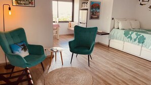 Ein Zimmer mit zwei smaragdgrünen Sesseln, einem kleinen runden Tisch, einem Bett mit einer grün-blumigen Tagesdecke und einem großen Fenster mit Blick auf Gebäude.