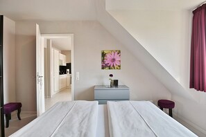 Habitación con dos camas, una mesita con un florero, un cuadro en la pared y vistas a una cocina y un baño por una puerta abierta.