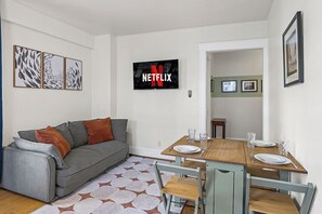 A living room with a grey sofa, a wooden dining table with chairs, a flat-screen TV, and framed pictures on the wall. - Charming Studio - Downtown Seattle - 98 Walk Score (Seattle)