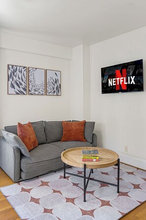 A living room with a gray sofa, a round coffee table with books, a patterned rug, and a wall-mounted Netflix sign. - Charming Studio - Downtown Seattle - 98 Walk Score (Seattle)