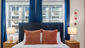 Een hotelkamer met een groot bed, twee nachtkastjes met lampen, een raam met jaloezieën en een muur met ingelijste foto's.
