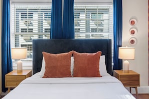 A hotel room with a large bed, two bedside tables with lamps, a window with blinds, and a wall with framed pictures.