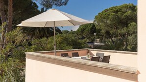 An outdoor patio with a table and chairs, a large umbrella, and a view of trees and a building in the background.
