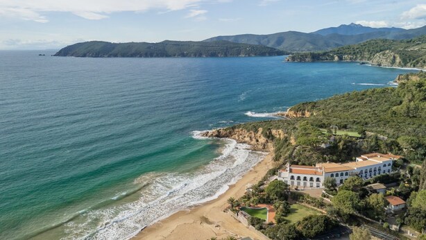 A coastal property with a beach, a large white building with arches, and surrounding greenery.