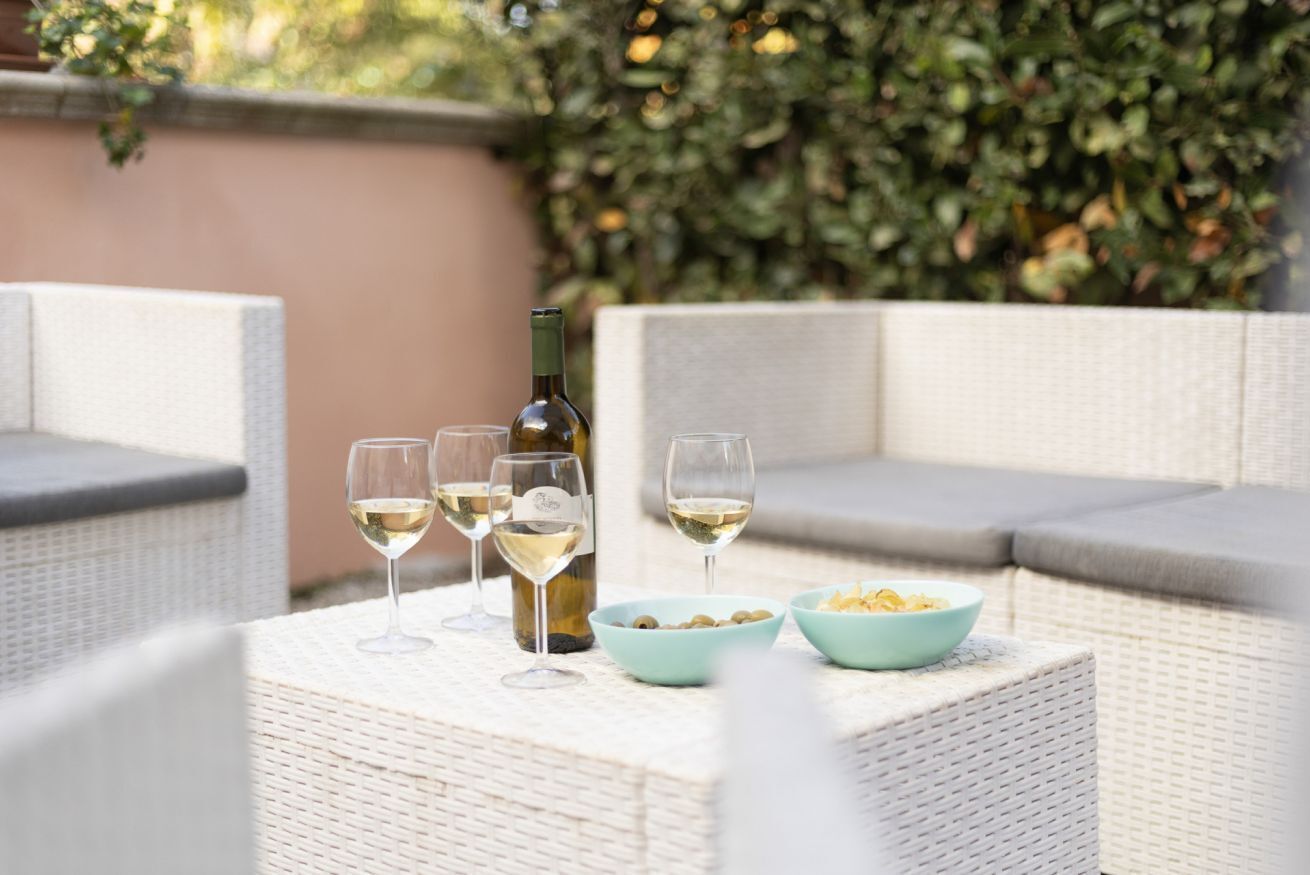 Une table en osier, avec une bouteille de vin, deux verres et des bols remplis de collations, sur une terrasse dotée d’un coin salon confortable et d’une haie verdoyante en arrière-plan.