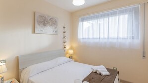 Ein Hotelzimmer mit einem Bett, einem Nachttisch mit Büchern, einer Lampe, einem Fenster mit durchsichtigen Vorhängen und einem Wandbild.