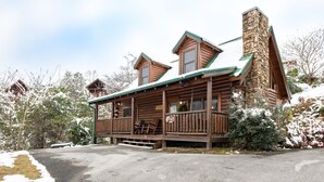 A log cabin with a stone chimney, wooden deck, and a snow-covered landscape. - Bearadise City🐻 Loft Cabin w/ Mtn Views, Hot Tub, & Pool Table! (Pigeon Forge)