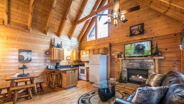 A cozy cabin interior with wooden walls, a fireplace, a TV, and a leather couch. - Bearadise City🐻 Loft Cabin w/ Mtn Views, Hot Tub, & Pool Table! (Pigeon Forge)