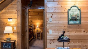 A cabin interior with a wooden wall, a welcome sign, a lamp, a small table with a decorative item, and a tray with mugs and a teapot. - Bearadise City🐻 Loft Cabin w/ Mtn Views, Hot Tub, & Pool Table! (Pigeon Forge)