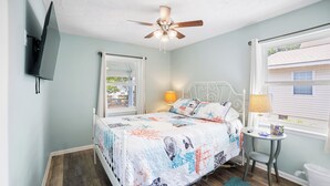 A bedroom with a bed, two bedside lamps, a ceiling fan, a window, and a small table. - The Inkwell ~ An Oak Island Gem (Oak Island)