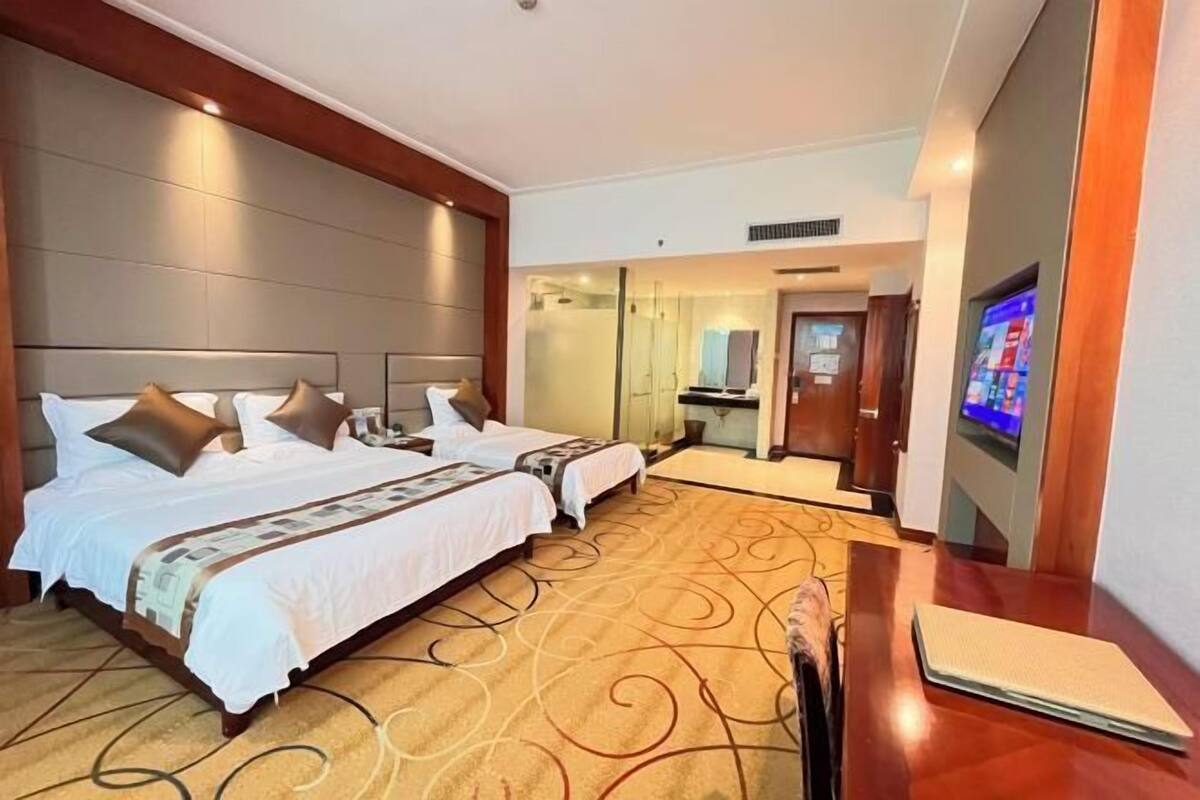 A hotel room with two beds, a TV, a desk, and a bathroom visible through a glass door.
