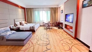Ein Hotelzimmer mit zwei Betten, einem Sofa, einem Esstisch, Stühlen, einem Fernseher und einem großen Fenster mit Vorhängen.