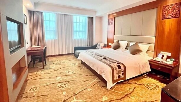 Ein Hotelzimmer mit einem großen Bett, einem Schreibtisch und einem Stuhl. Vor den Fenstern hängen Vorhänge und an der rechten Seite befindet sich eine dekorative Wandverkleidung.