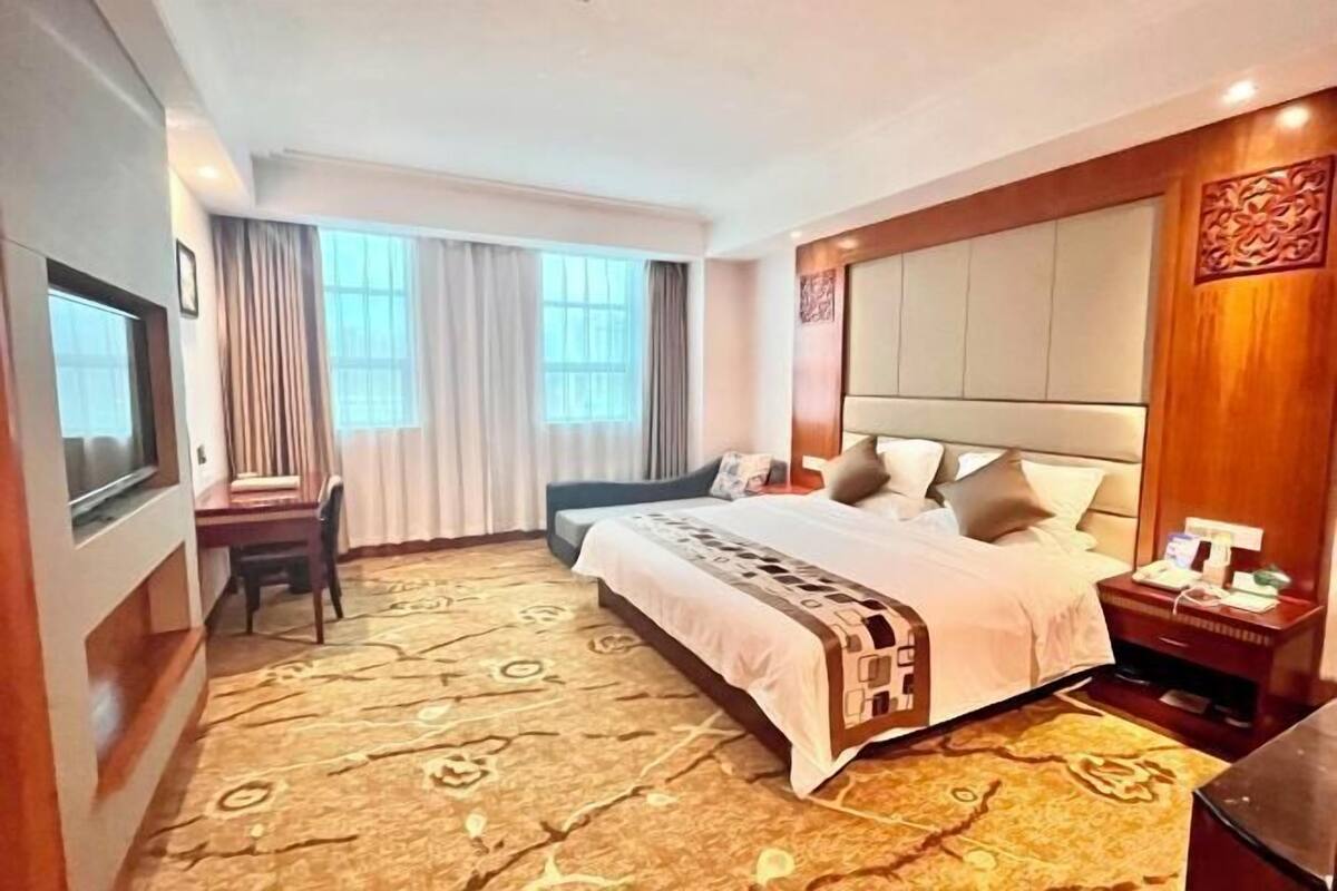 A hotel room with a large bed, a desk, and a chair. There are curtains on the windows, and a decorative wall panel on the right side.