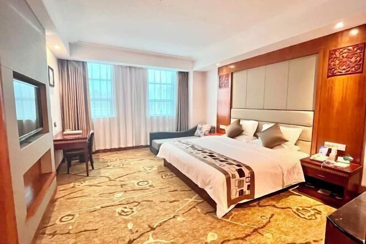 A hotel room with a large bed, a desk, and a chair. There are curtains on the windows, and a decorative wall panel on the right side.