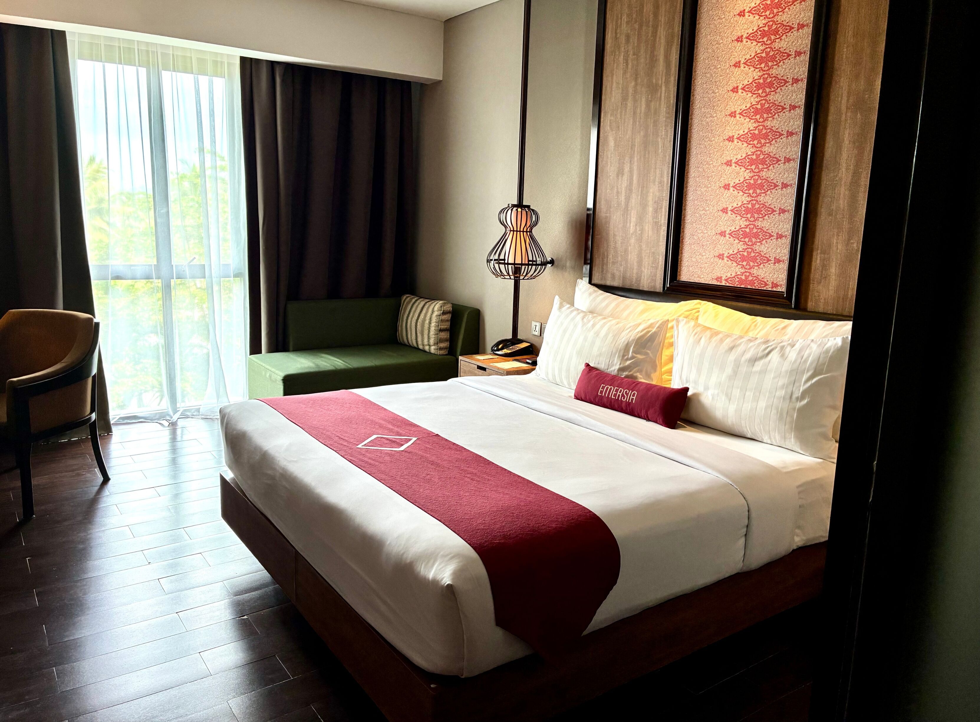 A hotel room with a large bed, a green sofa, a desk with a lamp, and a view of greenery through the window.