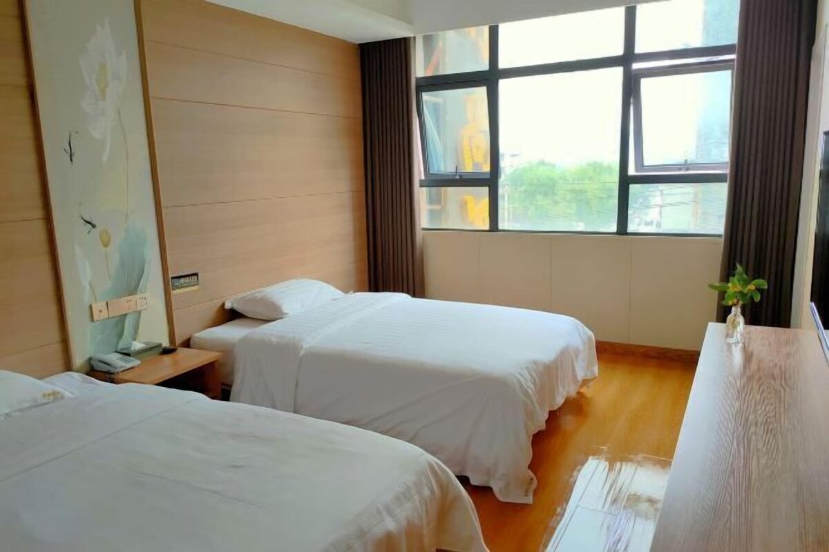 Een hotelkamer met twee bedden, een houten dressoir, een groot raam met gordijnen en een wand met een bloemschilderij.