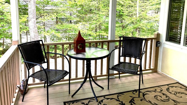 A covered porch with two black wicker chairs and a small table, overlooking a lush green landscape. - Point Sebago 3 BR, 1.5 bath Maine lake cottage near beach on Purple Rd (Casco)