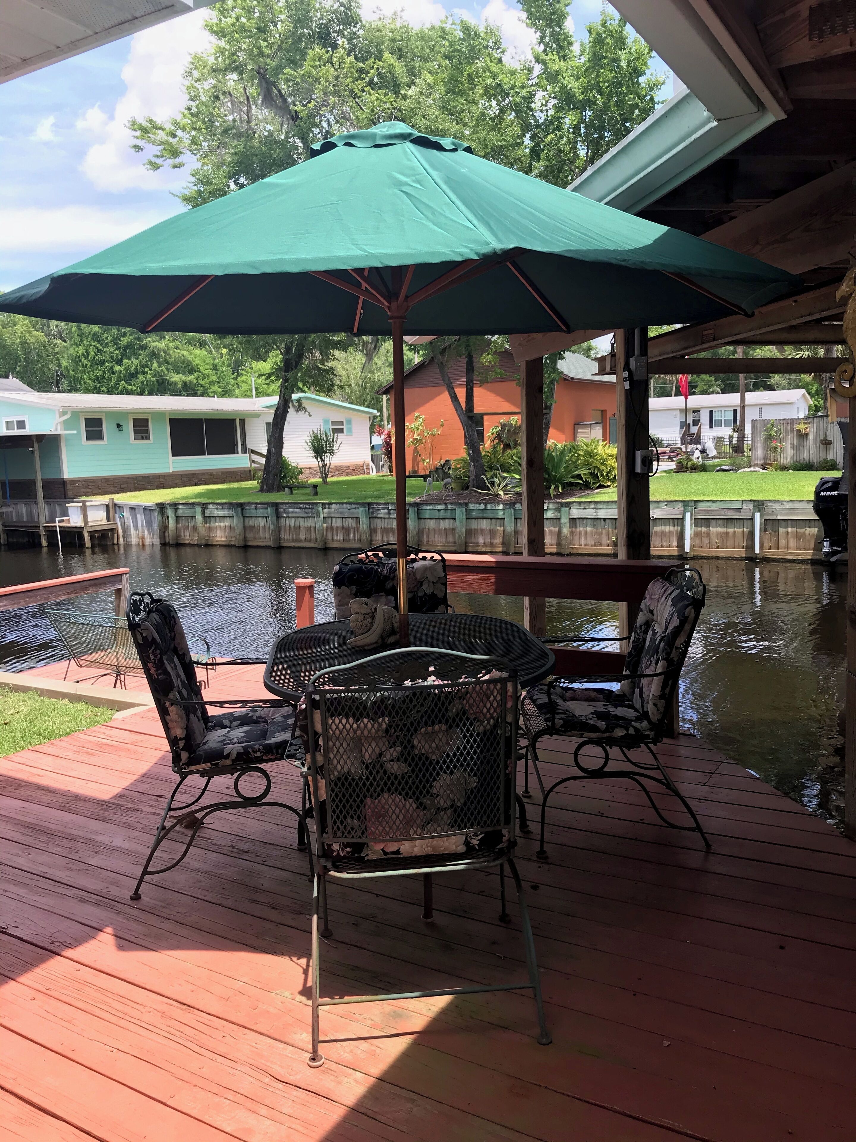 Une terrasse extérieure couverte, avec une table et des chaises, un parasol vert, et une vue sur une maison et un étang.